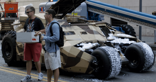 Two men stand near a camouflaged vehicle with snow on its tires, set against an urban backdrop.