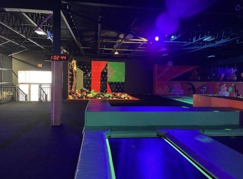 A brightly lit indoor trampoline park featuring trampolines, a climbing wall, and colorful foam pits.