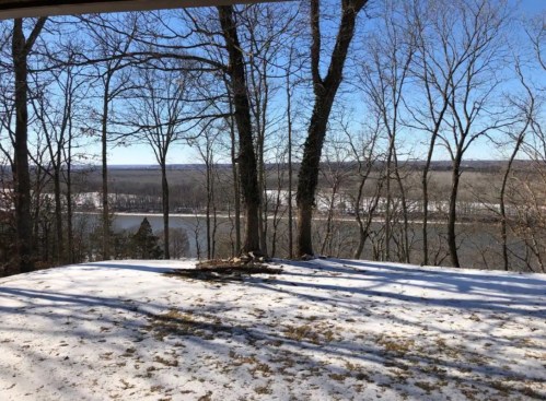 A snowy landscape with bare trees overlooking a river and distant hills under a clear blue sky.
