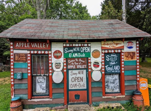A colorful, rustic building with signs indicating hours and attractions, surrounded by greenery.