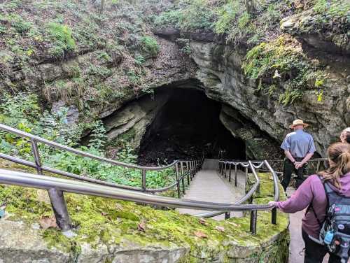 A group of people stands at the entrance of a cave, with stone steps leading down into the dark opening.