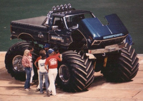 A large blue monster truck with oversized tires, surrounded by a group of people admiring it at an indoor event.