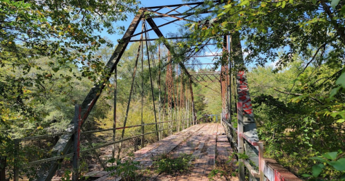 An old, rusted bridge surrounded by greenery, with wooden planks and graffiti visible on the structure.