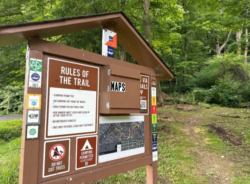 Trailhead sign displaying rules, maps, and information about camping and trail usage in a wooded area.