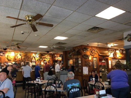 A busy diner interior with patrons at tables and a counter, featuring rustic decor and a ceiling fan.