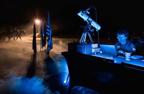 A person works at a desk with a telescope, surrounded by fog and illuminated by blue light, with horses and flags in the background.