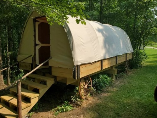 A wooden covered wagon with a canvas top, set in a wooded area with steps leading up to the entrance.