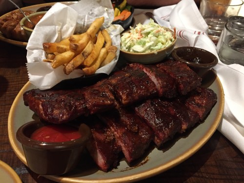 A plate of barbecue ribs with fries, coleslaw, and dipping sauces on a wooden table.