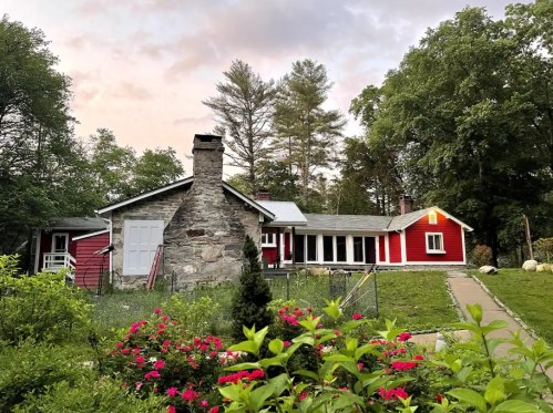 A charming red house with a stone chimney, surrounded by greenery and colorful flowers under a cloudy sky.
