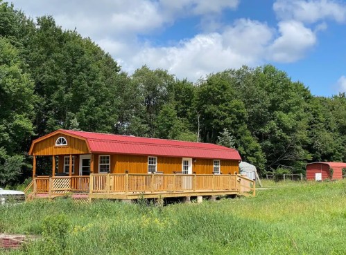 A bright orange cabin with a red roof sits on a wooden deck, surrounded by green grass and trees under a blue sky.