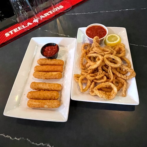 Two plates of appetizers: one with breaded mozzarella sticks and the other with fried calamari, served with marinara sauce.