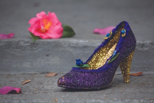 A sparkling purple high heel shoe adorned with flowers, resting on stone steps beside pink petals and a flower.