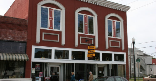 A red brick building with large windows and a sign reading "Open" at the entrance, featuring people walking by.