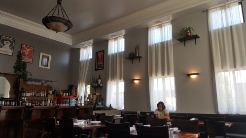 A cozy restaurant interior with large windows, plants, and a woman sitting at a table.