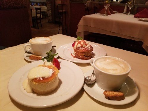 A table setting with two cups of coffee and three desserts: cheesecake, a fruit tart, and a cake with caramel drizzle.
