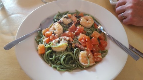 A plate of green pasta with shrimp, tomatoes, and herbs, garnished and served with utensils on a yellow tablecloth.