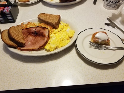 A breakfast plate with scrambled eggs, ham, hash browns, and toast, alongside a slice of cake with icing on a separate plate.