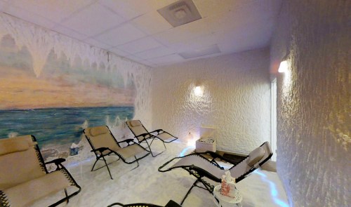 A serene spa room with reclining chairs, salt-covered walls, and a calming mural of the ocean. Soft lighting enhances relaxation.