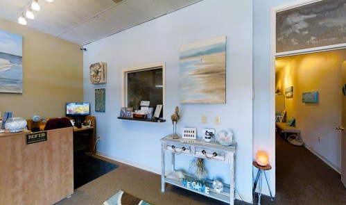 A cozy office reception area with artwork, a desk, and a hallway leading to another room. Soft lighting and decor present.