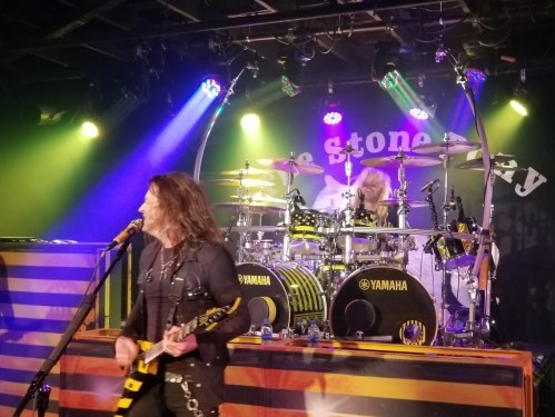 A rock band performs on stage with vibrant lights; a guitarist sings while a drummer plays in the background.