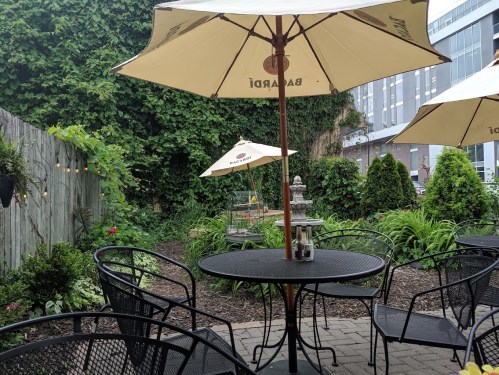 A cozy outdoor seating area with black tables and umbrellas, surrounded by lush greenery and plants.