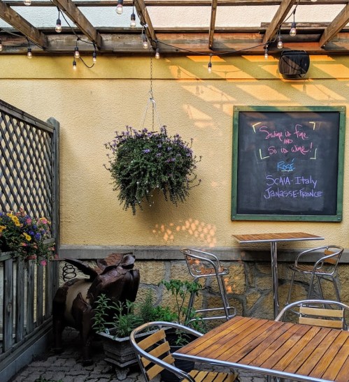 A cozy outdoor seating area with a hanging plant, wooden tables, and a chalkboard menu against a yellow wall.