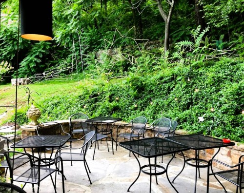 A serene outdoor café area with black metal tables and chairs surrounded by lush greenery and stone pathways.