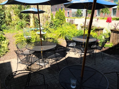 A sunny outdoor patio with black metal tables and chairs, shaded by blue umbrellas, surrounded by greenery.