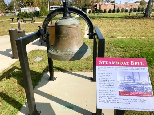 A large steamboat bell displayed outdoors, with an informational sign about its history and significance nearby.