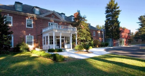 A large brick building with a porch, surrounded by greenery and trees, set in a sunny outdoor area.