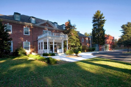 A brick building with a porch, surrounded by greenery and trees, on a sunny day.