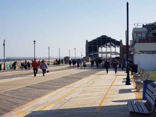 A busy boardwalk with people walking, benches, and a beach in the background under a clear blue sky.