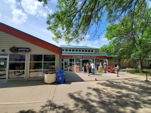 Entrance to Blank Park Zoo with visitors, surrounded by trees and a blue sky.