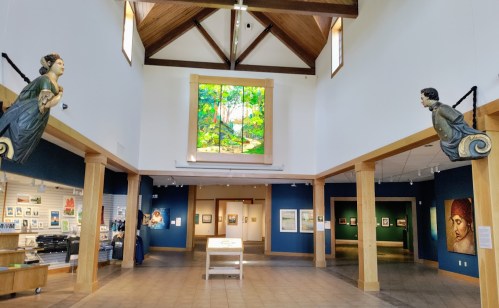 Interior of an art gallery featuring a large stained glass window, wooden beams, and sculptures on either side.