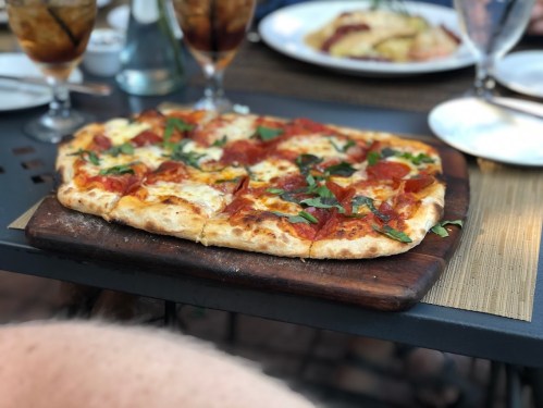A freshly baked pepperoni pizza topped with basil, served on a wooden board at a dining table.