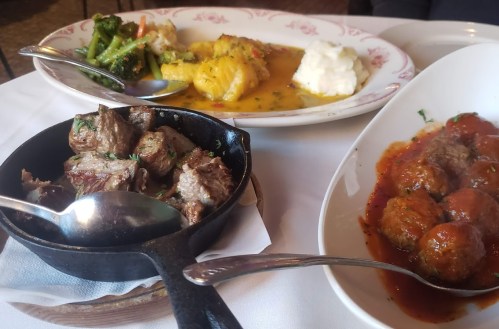 A table setting with three dishes: sautéed vegetables, mashed potatoes, and meatballs in sauce, alongside grilled meat.