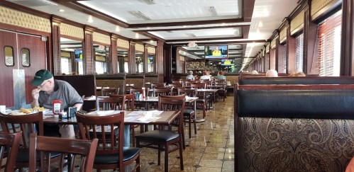 A spacious diner interior with booths and tables, featuring patrons dining and TVs in the background.