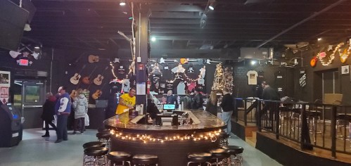 A lively bar scene with a wooden counter, patrons mingling, and walls decorated with guitars and memorabilia.