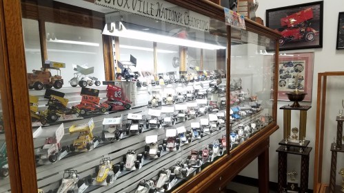 A display case filled with various miniature race cars and trophies in a museum setting.