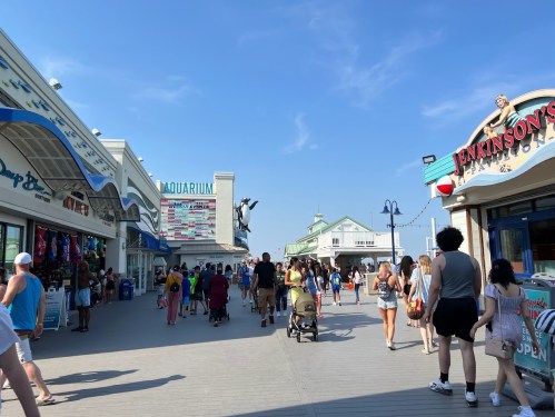 A busy boardwalk with shops and people walking, under a clear blue sky. An aquarium sign is visible in the background.