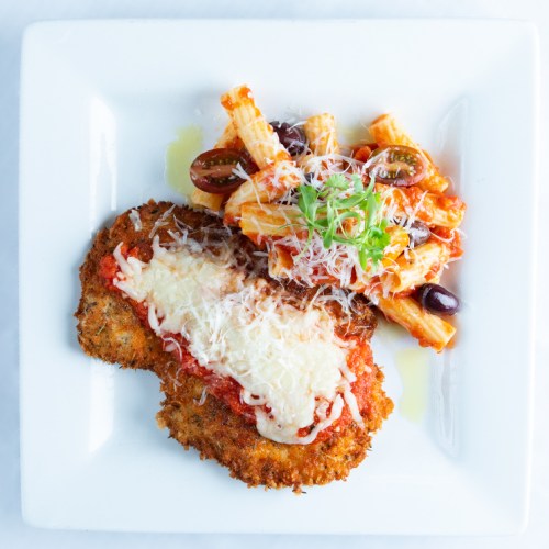 A plate featuring breaded chicken topped with marinara sauce and melted cheese, served with rigatoni pasta and olives.