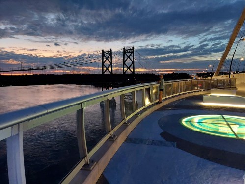 A person stands on a bridge at sunset, with a river and a bridge in the background under a colorful sky.