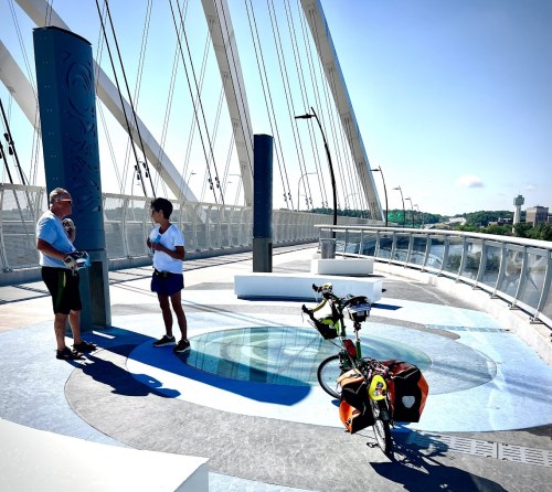 Two people converse on a bridge, with a bicycle and colorful panniers nearby, under a clear blue sky.
