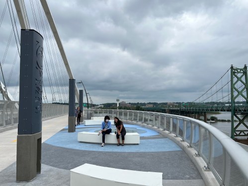 Two people sit on a curved bench on a bridge, with a cloudy sky and river in the background.