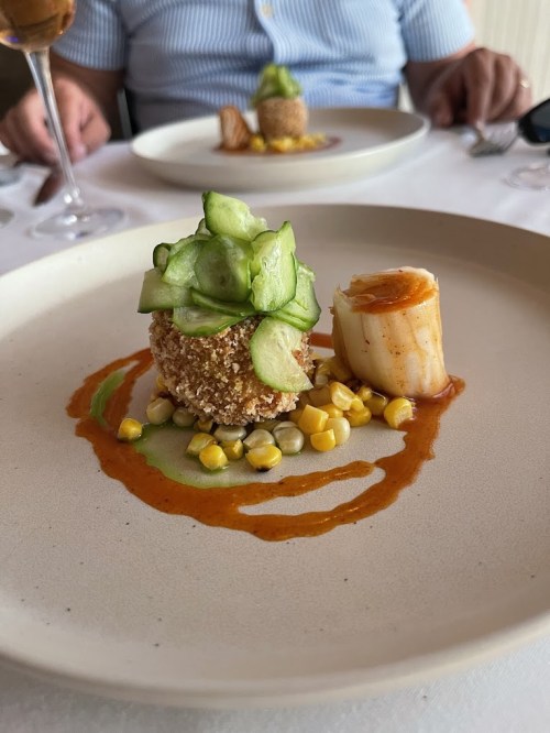 A gourmet dish featuring a breaded ball, scallop, corn, and cucumber on a plate with sauce drizzles.