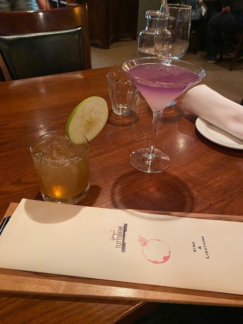 A wooden table with a cocktail menu, a green apple cocktail, and a purple martini in elegant glasses.
