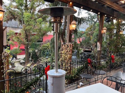 Outdoor dining area with a patio heater, surrounded by greenery and a small water feature, decorated for the holidays.