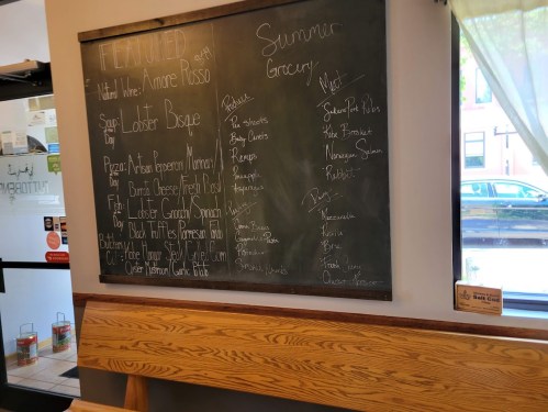Chalkboard menu featuring lobster bisque, summer grocery items, and various dishes listed in a cozy café setting.