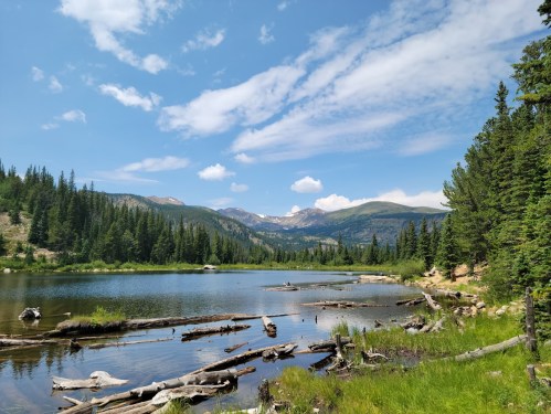 A serene lake surrounded by lush trees and mountains under a blue sky with scattered clouds.