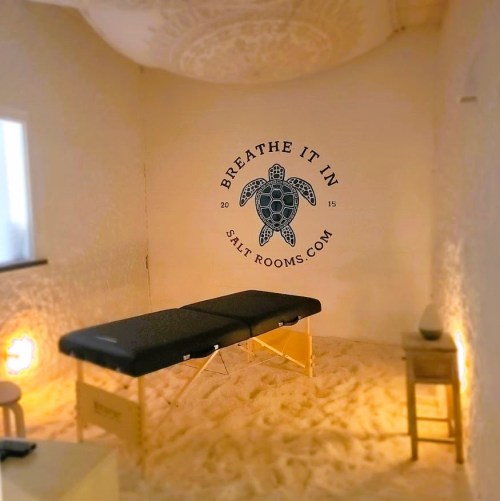 A serene salt room with a massage table, soft lighting, and a turtle logo on the wall, promoting relaxation.